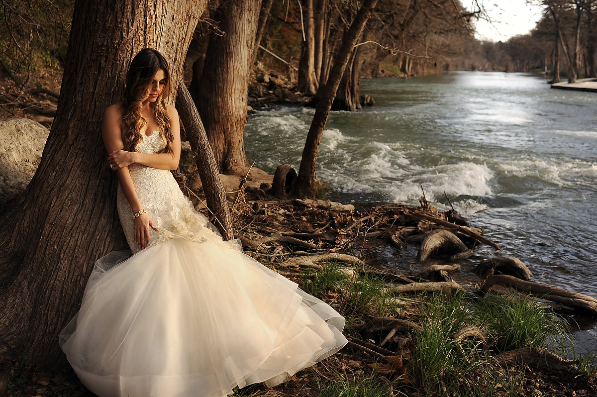 A bride is leaning against a tree at the side of the guadalupe river in gruene tx